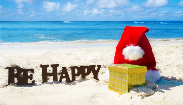 Sign "Be Happy" with christmas hat and gift box on the sandy beach by the ocean in sunny day