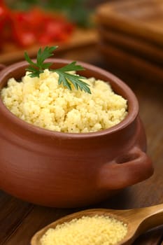 Prepared couscous in rustic bowl garnished with parsley, raw couscous on wooden spoon in front (Selective Focus, Focus on the front of the parsley leaf on the couscous)