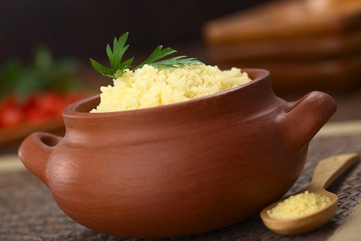 Prepared couscous in rustic bowl garnished with parsley, raw couscous on wooden spoon on the side (Selective Focus, Focus on the front of the parsley leaf on the couscous)