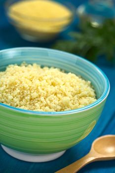 Prepared couscous in colorful bowl on blue wooden surface with raw couscous, parsley and oil in the back (Selective Focus, Focus one third into the couscous)