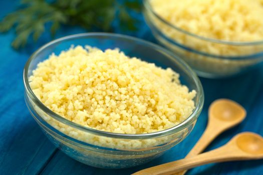 Prepared couscous in glass bowl on blue wooden surface with parsley in the back (Selective Focus, Focus one third into the couscous)