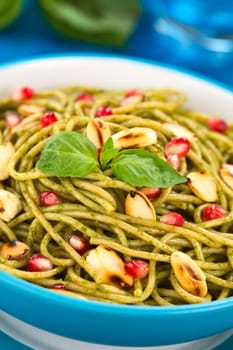 Spaghetti with pesto, pomegranate and roasted almond halves garnished with a basil leaf and served in a blue bowl on blue wood, with glass of water and basil leaf in the back (Selective Focus, Focus on the front of the basil leaf on the dish)