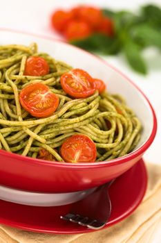 Spaghetti with pesto and baked cherry tomato halves served in a red bowl with a fork beside on white wood, with cherry tomatoes and basil leaf in the back (Selective Focus, Focus one third into the dish)