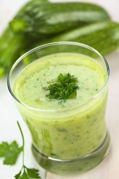 Zucchini cream soup garnished with curled parsley, served in glass on white wood with small zucchini in the back (Selective Focus, Focus on the front of the parsley on the soup)