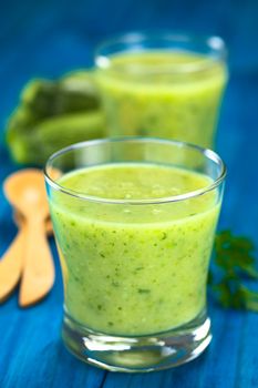 Zucchini cream soup served in glasses on blue wood with wooden spoon, parsley  and small zucchini in the back (Selective Focus, Focus on the front of the rim of the first glass)