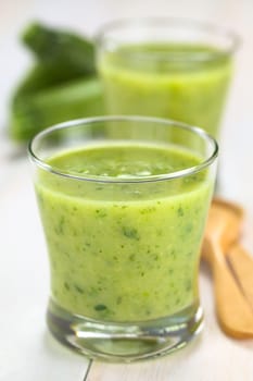 Zucchini cream soup served in glasses on white wood with wooden spoon on the right and small zucchini in the back (Selective Focus, Focus on the front of the rim of the first glass)