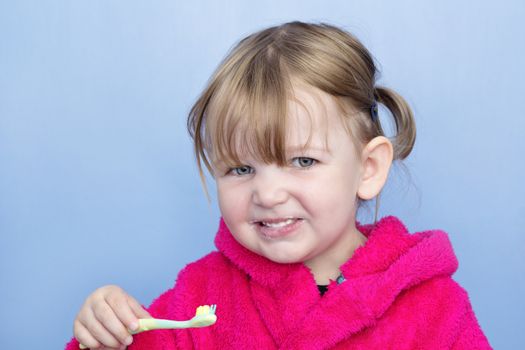 A young girl in a pink dressing gown against a light blue background. She's cleaning her teeth and smiling at the camera.