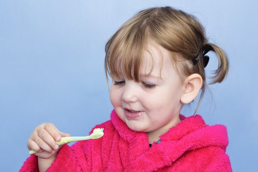 A young girl in a pink dressing gown against a light blue background. She's cleaning her teeth and has stopped to smile at the brush.