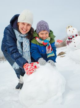 Happy beautiful boy with mother building snowman outside in winter time