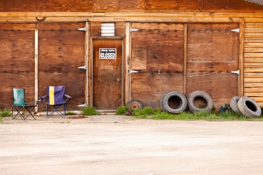 Wooden gate of rural timber building with closed sign and abandoned with two vacant chairs and old tires leaning against outside wall