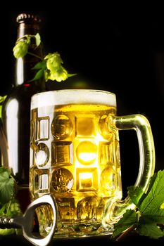 Mug of golden beer, bottle and openner with hop leaves over black