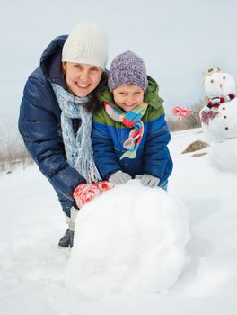 Happy beautiful boy with mother building snowman outside in winter time