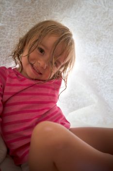 Playful cute Caucasian little girl wearing a pink striped T-shirt under a white blanket, shot from low angle