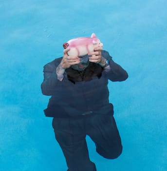Senior caucasian man holding piggy bank above water as he slowly drowns in debt wearing business suit