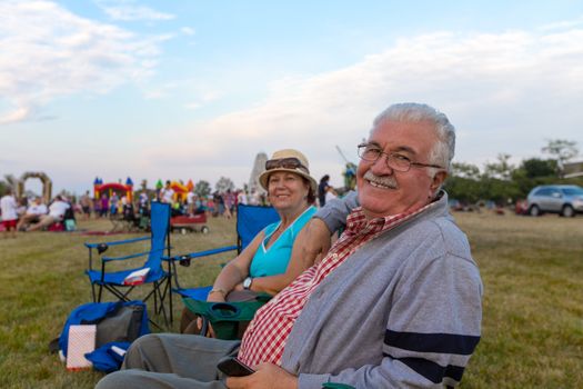 Elderly couple of spectators sitting in deckchairs at an outdoors event on a field turning to smile at the camera