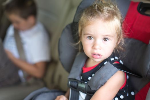 Cute little girl with a serious bewildered expression strapped into a childs safety seat in a vehicle with her young brother visible in the background