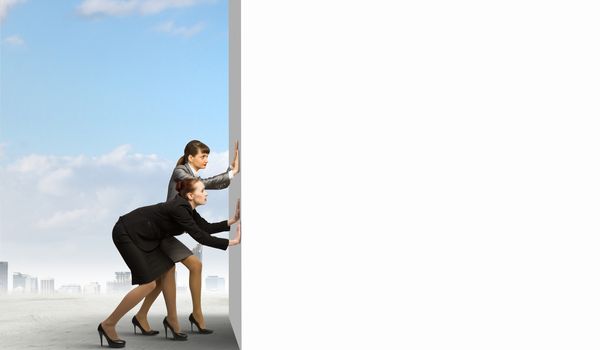 Image of two businesswomen pushing blank wall. Place for text