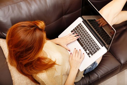 A young adult woman typing on a laptop while relaxing on the sofa.