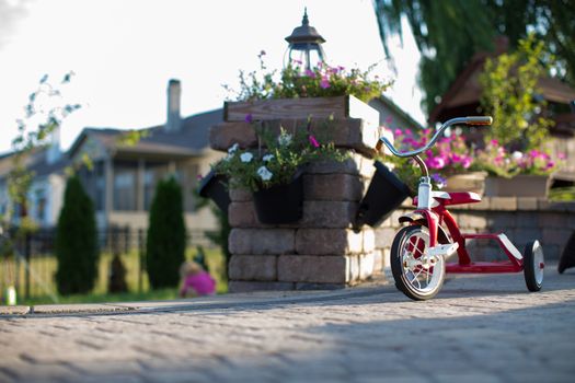 Childs red tricycle parked on a paved patio overlooking a garden in a shaft of sunlight