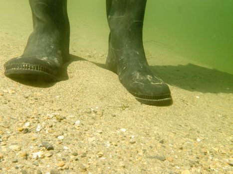 Closeup underwater view of rubber boots gumboots or waders of a person walking in shallow water of gravel and sand beach