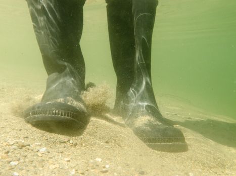Closeup underwater view of rubber boots gumboots or waders of a person walking in shallow water of gravel and sand beach
