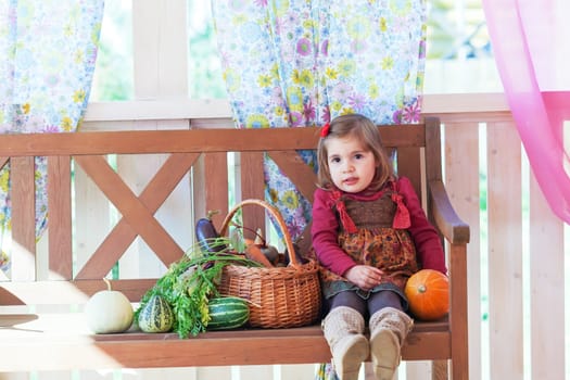 little girl sits on a bench on a terrace with a basket with vegetables