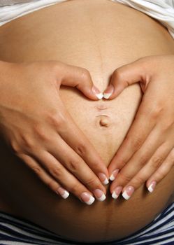 A woman who is eight months pregnant forms a heart over her belly with her hands.