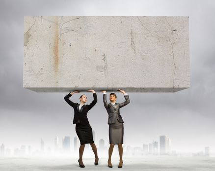 Image of two businesswomen holding stone above head. Partnership and cohesion