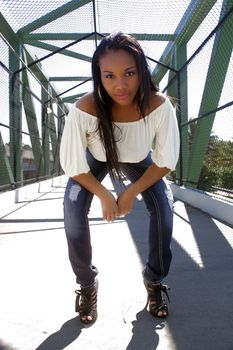 A lovely young Haitian girl on a pedestrian overpass covered with chain-link fence, bends over slightly toward the low-angle camera.