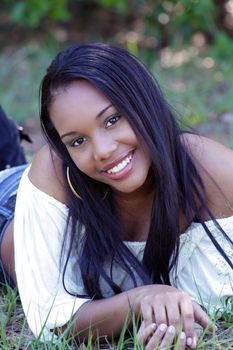 A close-up of a lovely young Haitian girl outdoors, lying in a grassy field, facing the camera, with a captivating smile.