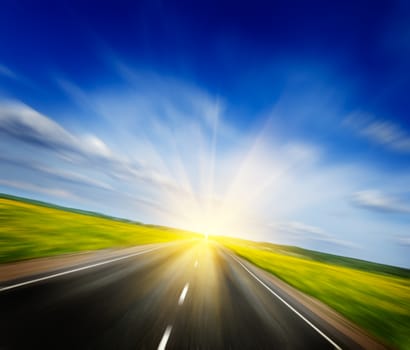 Travel concept background - motion blurred road in blooming spring meadow on sunset with blue sky