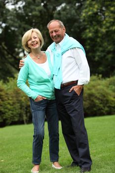 Laughing relaxed elderly couple standing arm in arm outdoors in the garden, full length portrait