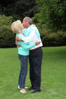 Romantic senior couple hugging each other and kissing as they stand close together in a green summer garden, full length portrait