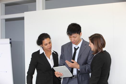 Three diverse young multiethnic business people having a team discussion standing looking at a tablet-pc in front of a presentation screen in the office