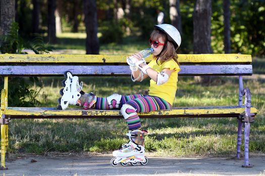 little girl with roller skates sitting on bench and drink water 
