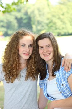 Two beautiful young teenage girls posing standing arm in arm outdoors and looking at the camera with warm friendly smiles