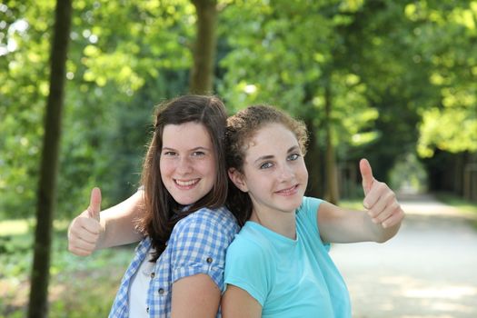 Happy attractive young teenage girls standing back to back in a park giving a thumbs up gesture of approval
