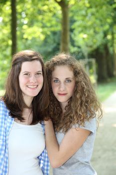 Two attractive young female friends in a park standing holding each other and smiling at the camera