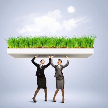 Image of two businesswomen holding lawn above head. Partnership and ecology