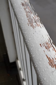 Flaking weathered white paint on a rain splattered wooden handrail.