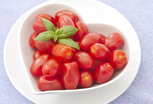 Fresh baby plum tomatoes in a heart shaped bowl, with a sprig of basil in the top left corner