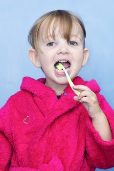 preschool girl cleaning her teeth. She's wearing a pink dressing gown against a blue background