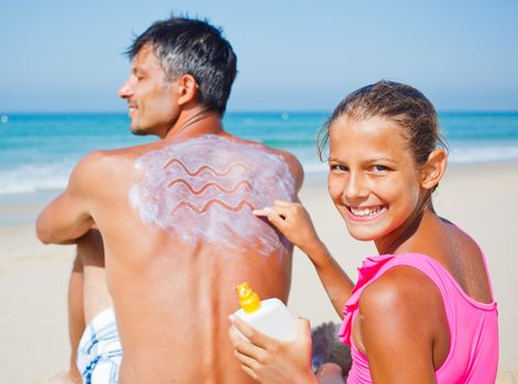 Adorable girl at tropical beach applying sunblock cream on a father's back.