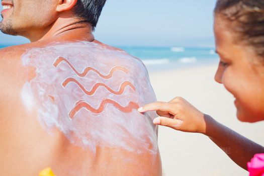 Adorable girl at tropical beach applying sunblock cream on a father's back.