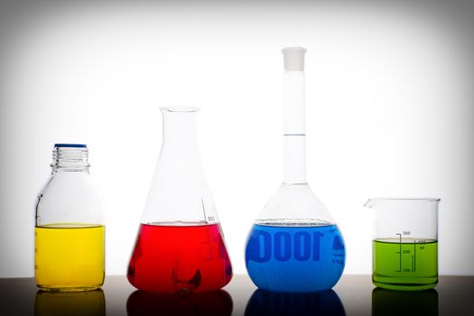 Four measuring glasses on a table of a chemical laboratory with red, yellow, blue and green liquid on white background