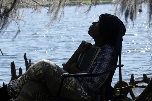 A lovely young woman looks away from her book at the waterfront, in silhouette.