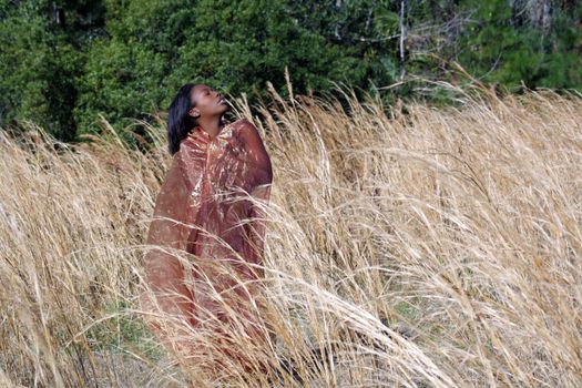 A lovely young black woman outdoors, standing in tall grass, wrapped in translucent fabric