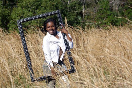 A lovely young black woman outdoors, standing in tall grass, holding a large black picture frame.  Generous copyspace on frame right.