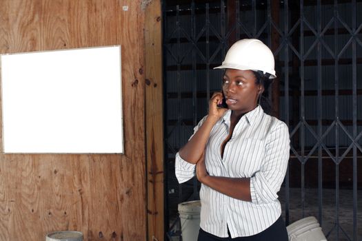 An attractive black woman wearing a white hardhat talks on her cell phone.  Beside and behind her is a white board on the wall where you can place your text or graphics.