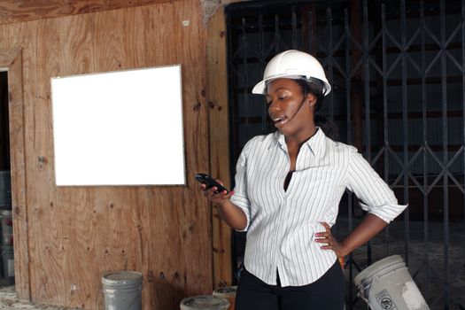 An attractive black woman wearing a white hardhat talks on her cell phone.  Beside and behind her is a white board on the wall where you can place your text or graphics.